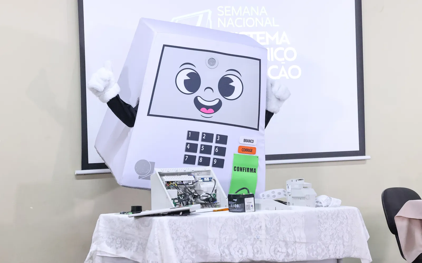 Mascot of the Brazilian electronic voting machine next to a ballot box and a projected slide